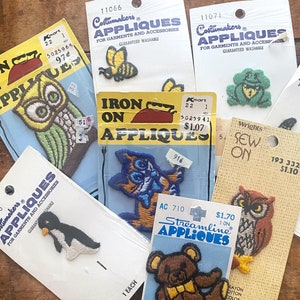 May include: A collection of iron-on appliques featuring various designs, including an owl, a penguin, a teddy bear, a frog, and a cat. The appliques are packaged on cards with prices and product information.