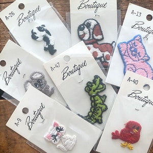 May include: Assortment of iron-on patches featuring various animal designs, including a cat, dog, bird, alligator, and koala. Each patch has a white background with black text that reads "Boutique" and a product code.