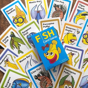 May include: A colorful Fish card game with a blue box featuring a smiling yellow fish. The cards are scattered, showcasing various sea creatures like the Tiger Shark, Ocean Sunfish, and Lion Fish. The game includes 37 cards.