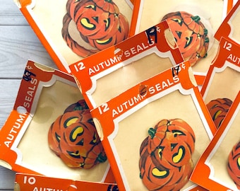 Könnte beinhalten: Eine Sammlung von 12 Vintage-Halloween-Aufklebern mit Kürbis-Designs. Die Blätter sind orange und weiß mit dem Text "12 Autumn Seals" und "Dennison" bedruckt.