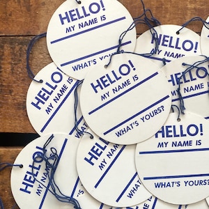 May include: A collection of white, circular name tags with blue text and string. Each tag reads "HELLO! MY NAME IS" and "WHAT'S YOURS?" in a bold, sans-serif font. The tags are arranged on a wooden surface.