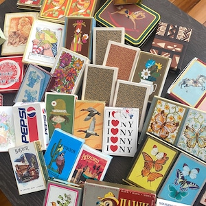 Puede incluir: Una gran colección de barajas de cartas vintage variadas con diversos diseños gráficos, incluyendo ilustraciones de mariposas, pájaros, flores, gatos, una rana, un caballito de madera y logotipos de Pepsi, I Love NY, Boston y American Airlines.