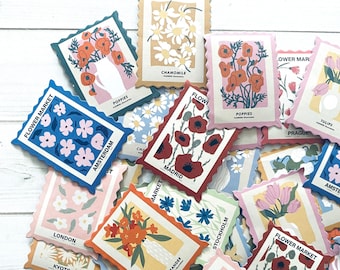 May include: A collection of colorful, illustrated stickers with flower designs and city names. The stickers are shaped like postage stamps and have a vintage aesthetic. The stickers feature flowers like poppies, chamomile, tulips, and oleander.