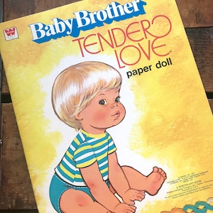 May include: A paper doll book featuring a baby boy with blonde hair and blue eyes. The book cover is yellow with the title "Baby Brother Tender Love Paper Doll" in red and blue lettering.