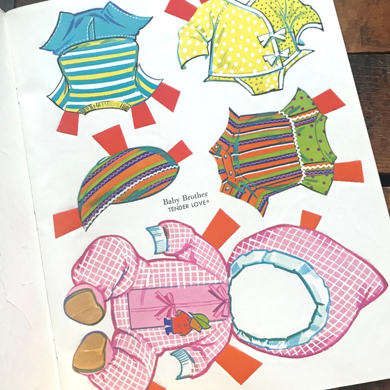 May include: A paper doll set with a baby doll and various outfits. The outfits include a pink and white checkered jumpsuit, a green and orange striped dress, a yellow and white polka dot dress, a blue and white striped shirt, and a blue hat. The text "Baby Brother Tender Love" is printed on the page.