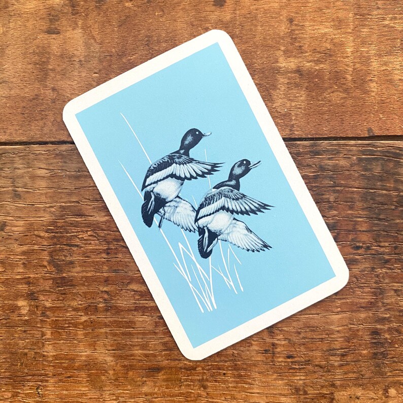 May include: A playing card featuring two ducks in flight, rendered in black and white against a light blue background. The card has a white border and is set against a wooden surface.
