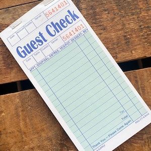 May include: A white and blue guest check with the words "Guest Check" in large blue letters. The check has a series of lines for itemizing orders and a space for the server's number. The check is on a wooden surface.