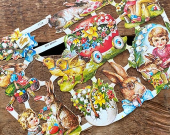 May include: A set of vintage Easter paper cutouts featuring bunnies, chicks, a basket of flowers, and a red wagon with flowers. The cutouts are printed on white paper with colorful illustrations. The text "ges. geschen." is printed on the paper.