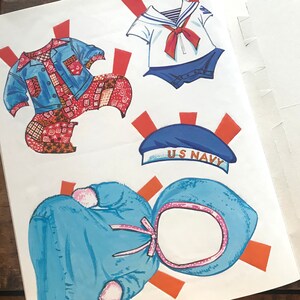 May include: Paper doll cutouts of a sailor, a patchwork dress, and a blue hooded dress. The sailor has a red and white striped shirt with a red bow tie and blue pants. The patchwork dress is red, white, and blue. The blue hooded dress has a pink trim. The sailor has a hat that says "US Navy".