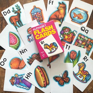 May include: A collection of alphabet flash cards with colorful illustrations and the letters of the alphabet. A pink box of flash cards is in the center, with the words "ALPHABET FLASH CARDS" printed on it. The cards feature images like a deer, fox, and butterfly.