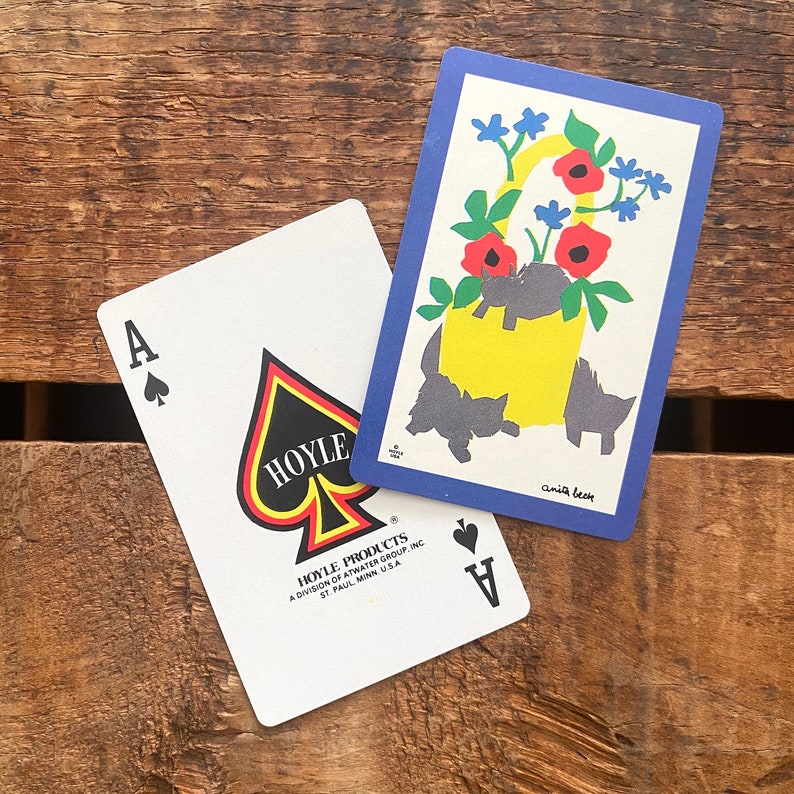Puede incluir: Dos cartas de juego, una es un as de picas con una pica negra y el texto "HOYLE" en ella. La otra carta es una carta azul con una cesta amarilla llena de flores y el texto "anita berse".