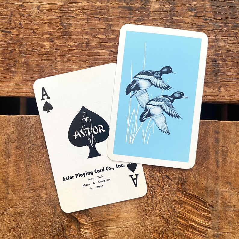 May include: Two playing cards featuring the Ace of Spades and the Ace of Diamonds. The Ace of Spades has a black spade design with the word "ASTOR" and a figure. The other card has a blue background with illustrations of ducks.