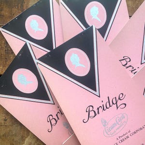 May include: Pink and black vintage packaging with a triangular design. Each package features a silver silhouette in a pink circle and the word "Bridge" in elegant script. The packaging also includes the text "Cream-Crest Milk Products".