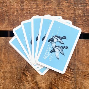 May include: A stack of playing cards with a light blue background and a black and white illustration of two ducks in flight. The cards are fanned out on a wooden surface.
