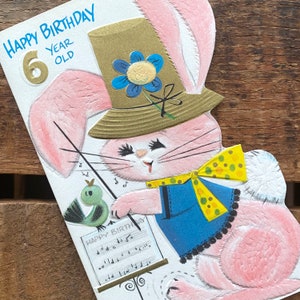 May include: A pink bunny rabbit wearing a brown hat with a blue flower and a yellow polka dot bow tie. The bunny is holding a music stand with a sheet of music that says "Happy Birthday". The bunny is holding a music stand with a sheet of music that says "Happy Birthday". The bunny is 6 years old.