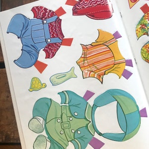 May include: A paper doll set with multiple outfits for a child. The outfits include a blue and red overalls, a yellow and orange striped shirt, a green jacket with a blue hat, and a red hat. The set also includes a small turtle and a whale.