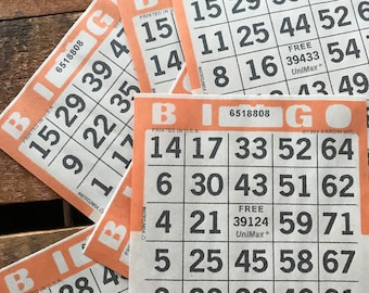 May include: Several orange and white bingo cards are arranged on a wooden surface. The cards feature the word "BINGO" at the top, along with numbered squares. The cards are printed in the USA.