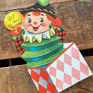 Puede incluir: Tarjeta de cumpleaños vintage con un payaso sonriente. El payaso tiene un cuello a rayas verdes y blancas y un sombrero negro con un pompón verde. La tarjeta presenta un diseño desplegable con un patrón de diamantes en rojo, blanco y rosa. El texto dice "Hi! It's Your Birthday."
