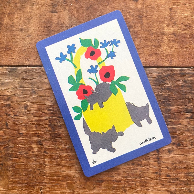 Puede incluir: Una tarjeta azul y blanca con una cesta amarilla llena de flores rojas y gatos negros. La tarjeta tiene un borde azul y el texto "Anita Beer" en la esquina inferior derecha.