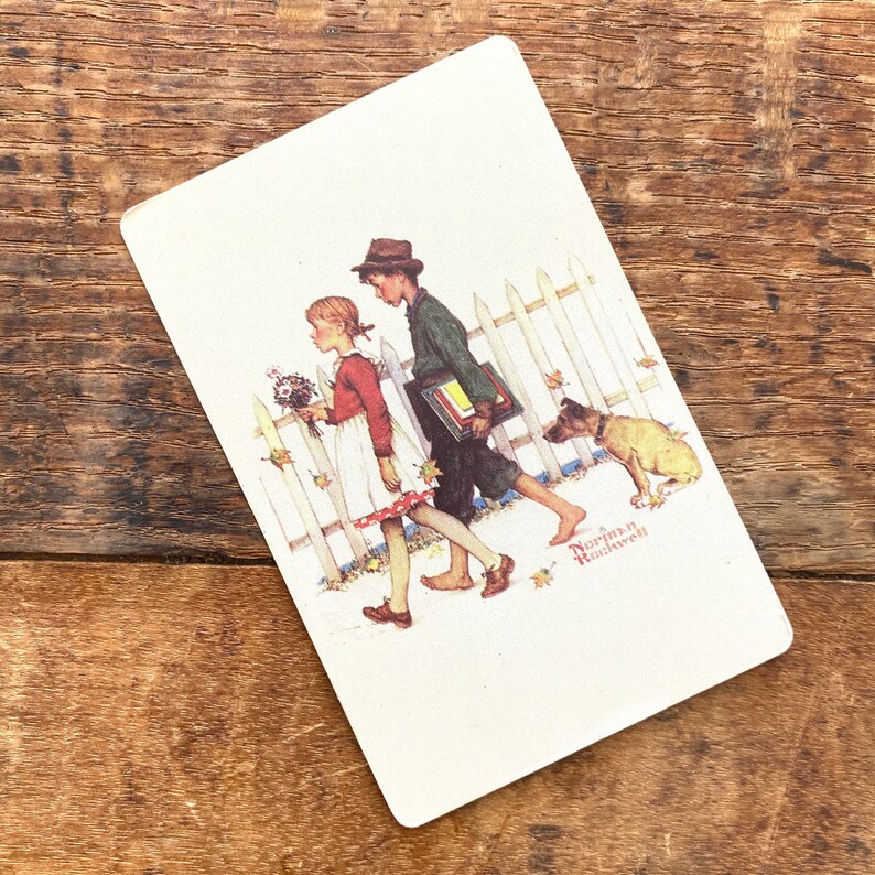 May include: A vintage playing card featuring a Norman Rockwell illustration. The image depicts a boy and girl walking past a white picket fence with a dog. The girl carries flowers, and the boy carries books. The card is set against a wooden background.
