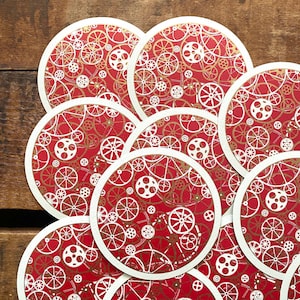May include: A collection of round coasters with a red background and a repeating pattern of white and gold gears. Each coaster has a white border. The coasters are arranged in a slightly overlapping pattern.