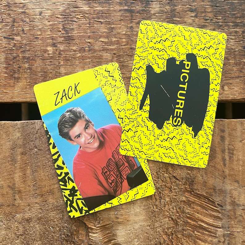 May include: Two yellow trading cards with black zig-zag patterns. One card features a photo of a smiling man in a red shirt with the name "ZACK" in green. The other card has the word "PICTURES" in black.