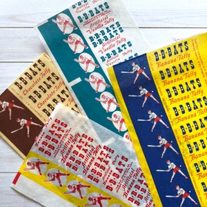May include: Vintage packaging for B-B-Bats banana taffy candy. The packaging features a blue and white design with a red, white, and blue baseball bat and a red, white, and blue baseball player. The text on the packaging reads "B-B-Bats Banana Taffy" and "Fair Play Caramels Inc., Johnson City, N.Y."