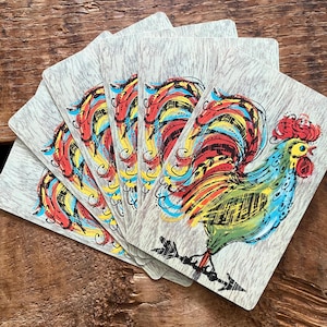 Puede incluir: Un juego de cartas con una ilustración de gallo colorido. El gallo tiene una cresta roja, un pico amarillo y una cola vibrante con plumas rojas, amarillas y azules. Las cartas tienen un fondo de vetas de madera gris claro.