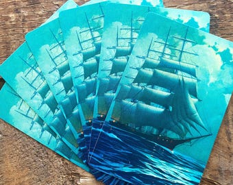 May include: A stack of rectangular cards featuring a vintage illustration of a tall ship sailing on a blue sea under a cloudy sky. The cards have a teal background and a rounded top edge. The ship is black with white sails.