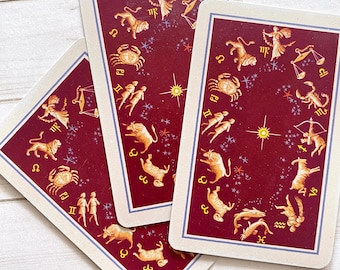 May include: Three rectangular tarot cards with a deep red background and a light blue border. The cards feature gold-colored illustrations of the twelve zodiac signs arranged in a circle, accompanied by small blue stars and a central gold star.