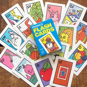 May include: A set of colorful phonics flash cards with illustrations and letters, along with a blue box of cards. The cards feature images like a swan, unicorn, and apple. The box reads "PHONICS FLASH CARDS" and "AGES 5 AND UP."