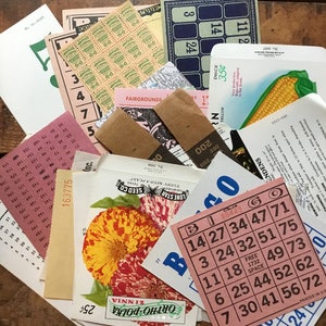 May include: A collection of vintage ephemera, including a bingo card, seed packets, a corn husk, and a ticket for a fairground. The bingo card is pink with black numbers and the words "Bingo" and "Go". The seed packets are brown with white text and a picture of a flower. The corn husk is yellow and brown. The ticket is pink with black text and the words "Fairgrounds".