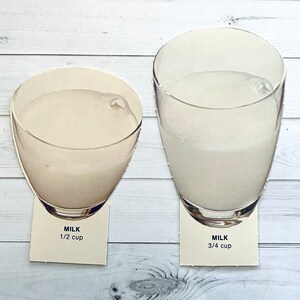 May include: Two glass cups filled with milk, one with 1/2 cup and the other with 3/4 cup of milk. The cups are on a white wooden surface.