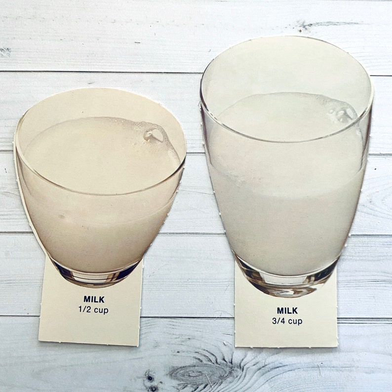 May include: Two glass cups filled with milk, one with 1/2 cup and the other with 3/4 cup of milk. The cups are on a white wooden surface.