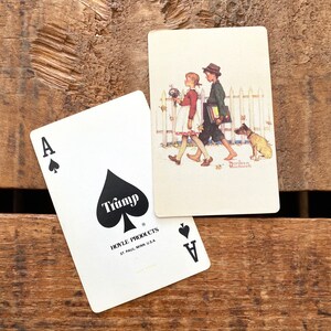 May include: Two playing cards on a wooden surface. One card is the Ace of Spades with the word "Trump" and "Hoyle Products" printed on it. The other card features a Norman Rockwell illustration of two children and a dog.