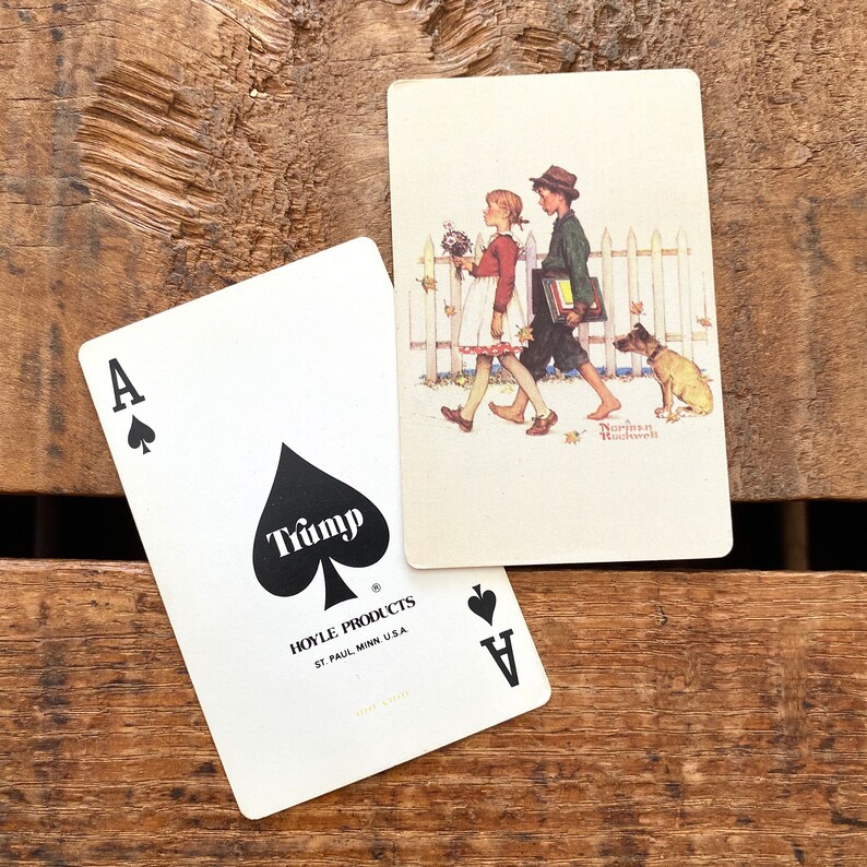 May include: Two playing cards on a wooden surface. One card is the Ace of Spades with the word "Trump" and "Hoyle Products" printed on it. The other card features a Norman Rockwell illustration of two children and a dog.