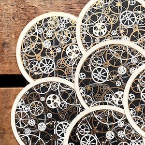 May include: Set of round coasters with a black background and a repeating pattern of gears in gold and white. The coasters have a cream-colored border and are arranged on a wooden surface. The design evokes a steampunk aesthetic.
