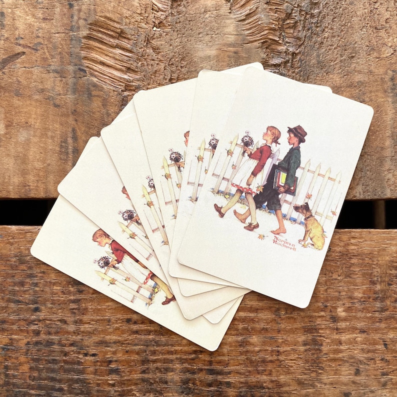 May include: A collection of playing cards featuring a vintage illustration of two children walking past a white picket fence with a dog. The artwork is signed "Norman Rockwell". The cards are arranged on a wooden surface.
