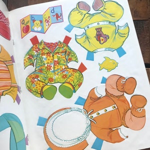 May include: A paper doll set with three different outfits. The outfits are a yellow and orange outfit with an elephant design, a green and yellow floral outfit, and an orange outfit with a white collar.