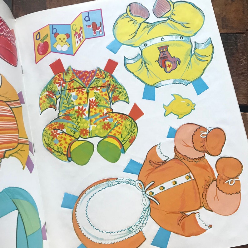 May include: A paper doll set with three different outfits. The outfits are a yellow and orange outfit with an elephant design, a green and yellow floral outfit, and an orange outfit with a white collar.