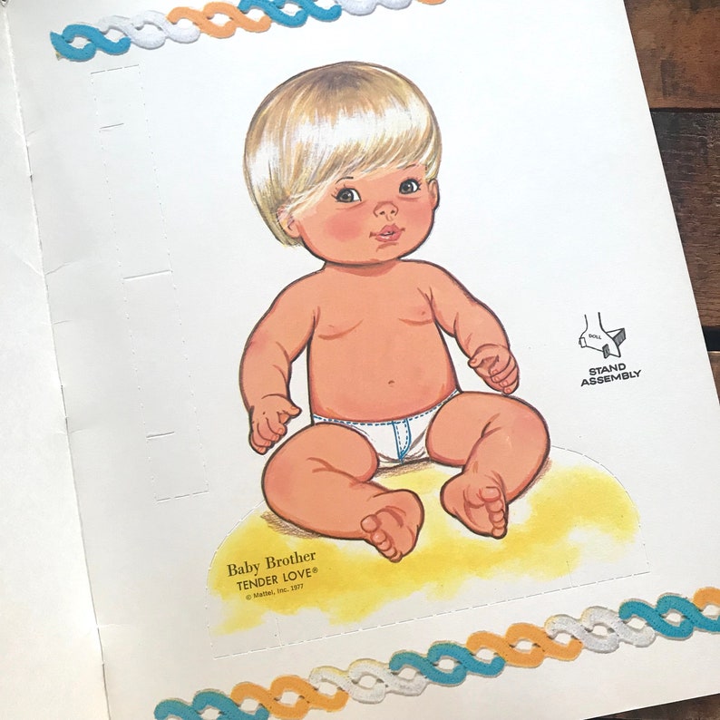 May include: A paper doll of a baby boy with blonde hair, wearing a white diaper and sitting on a yellow base. The doll is part of a paper doll set called "Baby Brother Tender Love".