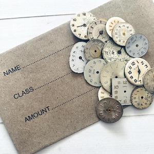 Puede incluir: Una colección de esferas de reloj vintage de varios tamaños y diseños, dispuestas sobre un fondo de papel marrón. Las esferas tienen esferas envejecidas de color blanquecino con números y manecillas negras. El papel tiene líneas para 'NOMBRE', 'CLASE' y 'CANTIDAD'.