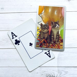 May include: A playing card with the Ace of Clubs printed on it. The card is lying on a white wooden surface. A photo of two kittens with balloons is visible in the background.