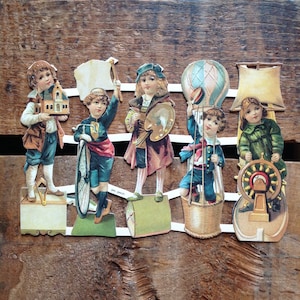 May include: Vintage paper cutouts of children in various occupations, including an architect, cyclist, artist, balloonist, and captain. The figures are dressed in period clothing and stand on decorative bases, with a wooden background.