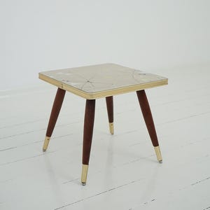 May include: A square side table featuring a light-colored top with a geometric pattern of thin black lines and small triangles. The table has four tapered dark wood legs, each finished with a metallic gold-toned cap at the base and a gold trim.
