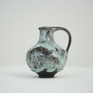 Può includere: Un piccolo brocca in ceramica verde e marrone con corpo arrotondato e manico. La brocca ha una smaltatura macchiettata.