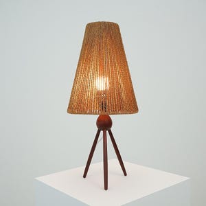 May include: A mid-century modern table lamp with a woven shade and a tripod base. The shade is a light brown color, and the base is dark brown. The lamp is approximately 24 inches tall.