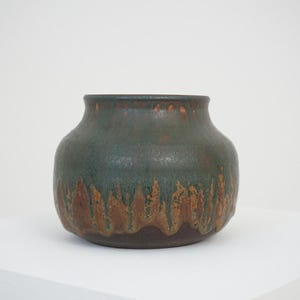 May include: A squat, rounded ceramic vase featuring a dark, matte brown base that transitions into a textured, mottled green glaze. The upper portion of the vessel displays a smooth, dark green finish with subtle drips of brown glaze running downward.