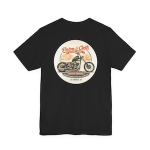 May include: Black t-shirt featuring a circular graphic with a motorcycle illustration. The graphic includes the text "Piston & Cloth Speed Shop, Parts & Service, Unauthorized Service & Repairs, Los Angeles, CA."