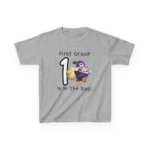 May include: A heather gray t-shirt with the text "First Grade" above a large number "1" and a cartoon character holding a bag. The text "Is In The Bag!" is below the character. The cartoon character is purple, yellow, and white.